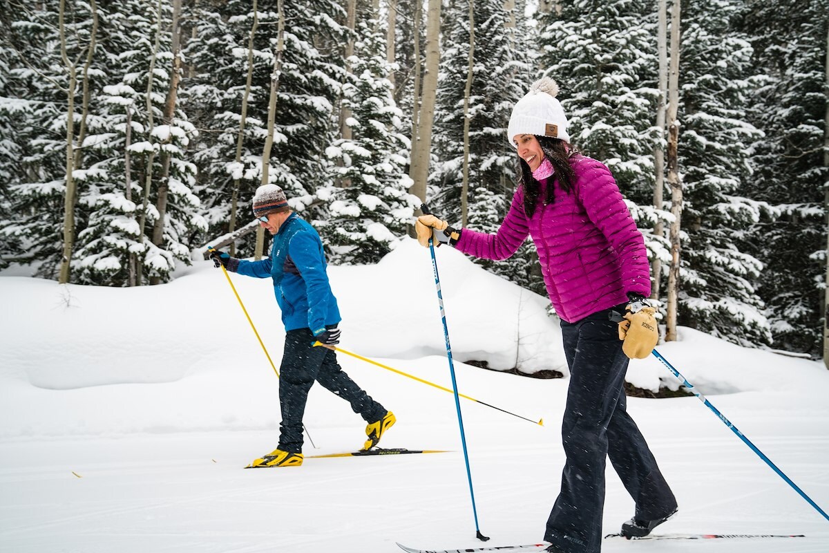 Two skiers gliding through deep snow on a groomed trail beneath snow-laden evergreens at Snow Mountain Ranch Nordic Center