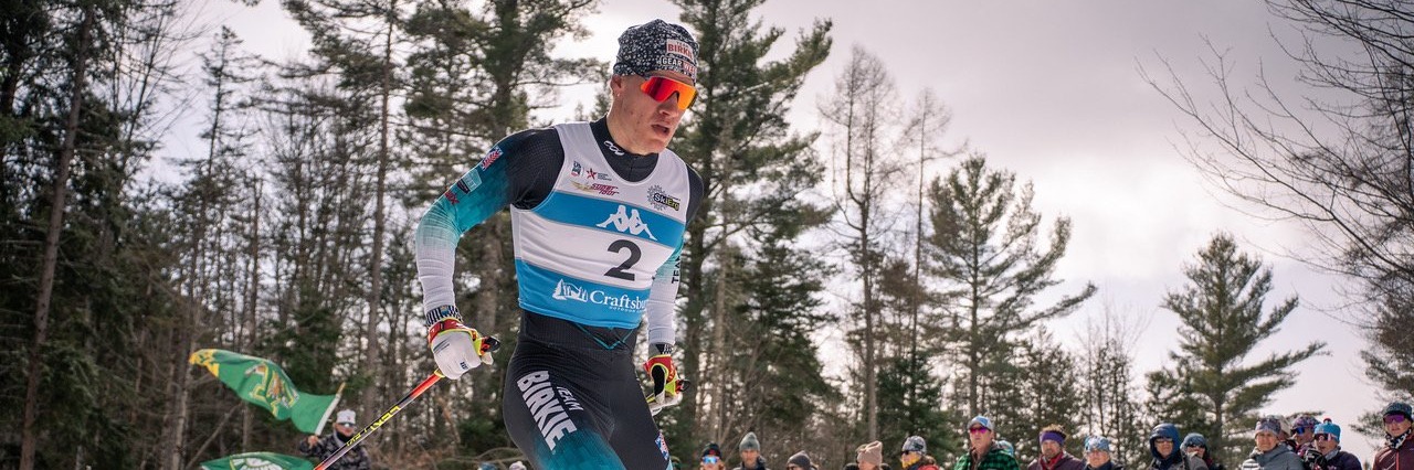 A Snø Racing skier striding past a line of teammates, coaches and families waving flags along the barrier at a winter race in Craftsbury, Vermont