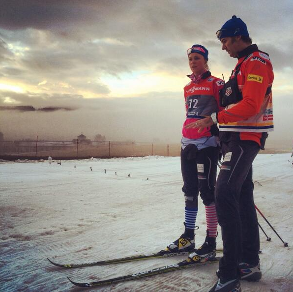 Randy Gibbs in a red jacket and blue cap stands on skis beside American athlete Sadie Bjornsen on a snow course at dusk